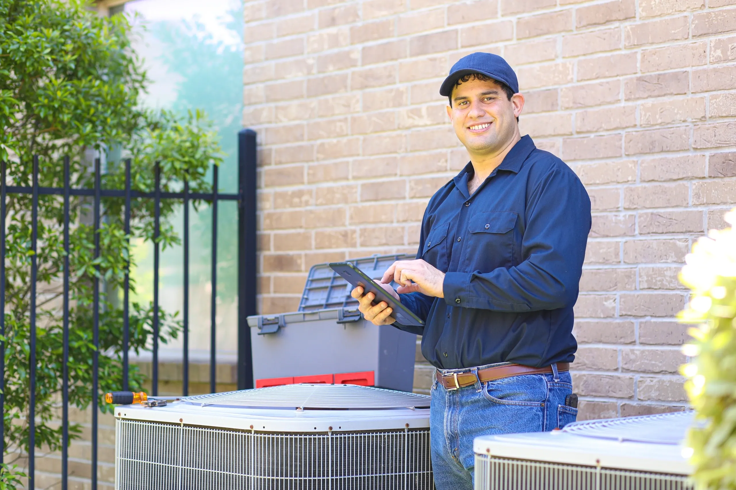 A smiling male technician in a cap stands next to an outdoor HVAC unit, holding a tablet.
