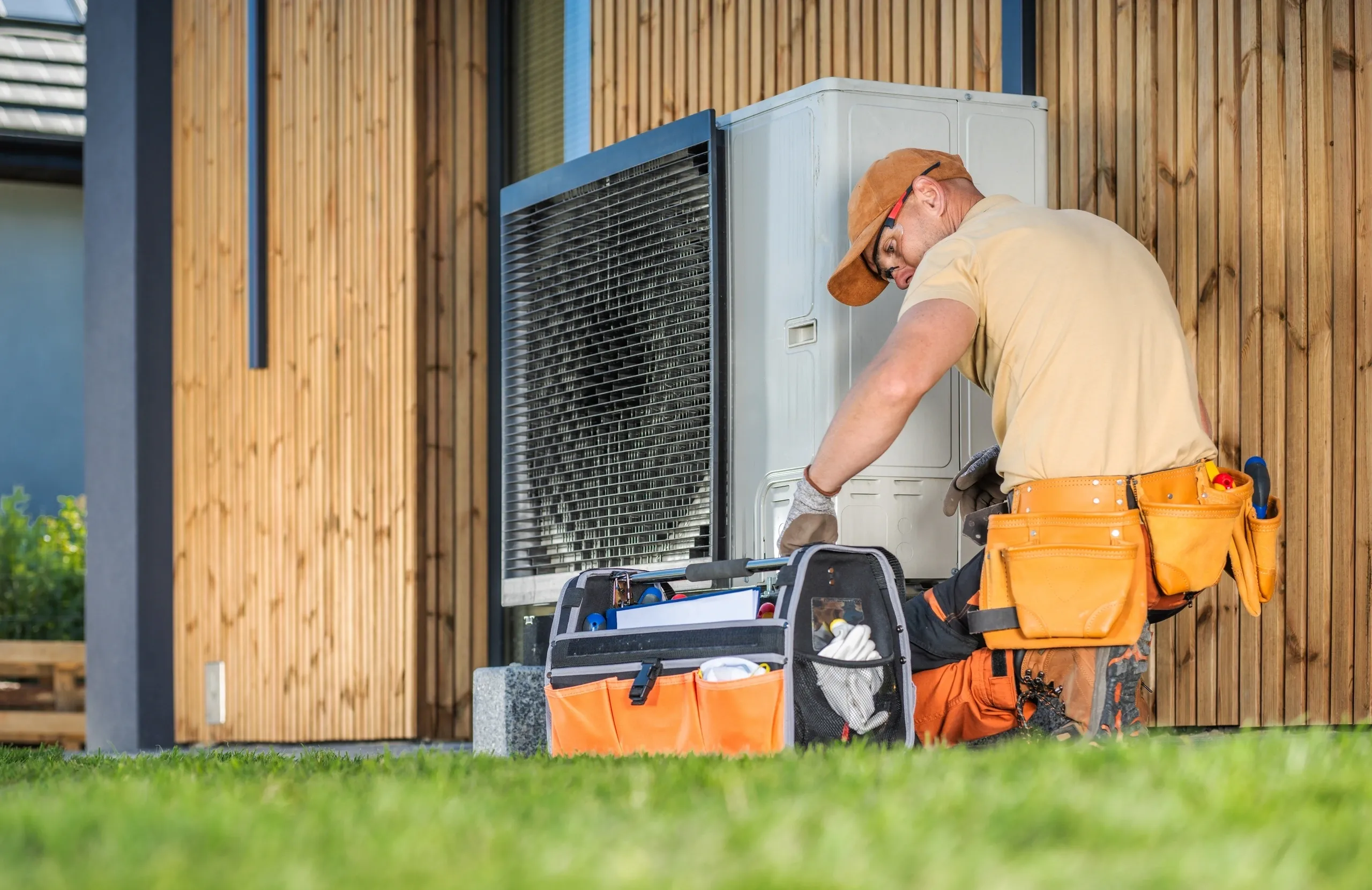 A male technician services an outdoor air conditioning unit next to a modern wooden house.